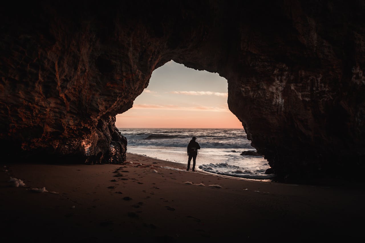 A person stands in a cave overlooking the ocean at sunset, creating a dramatic silhouette.