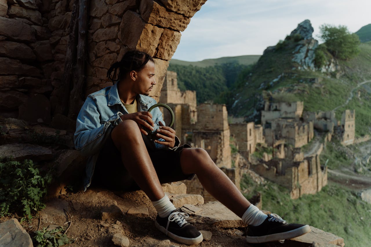 Young man in denim jacket with headphones, sitting amidst mountain ruins at sunset.